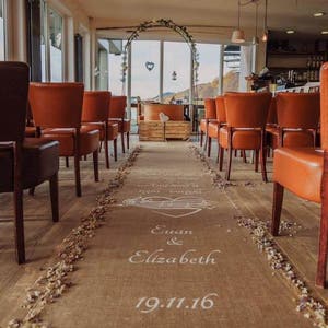 May include: A wedding ceremony aisle decorated with a burlap runner and white flowers. The aisle is lined with brown and orange chairs. The runner has the names "Euan & Elizabeth" and the date "19.11.16" written on it.