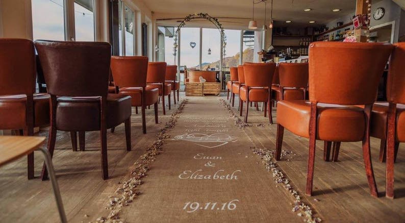 May include: A wedding ceremony aisle decorated with a burlap runner and white flowers. The aisle is lined with brown and orange chairs. The runner has the names "Euan & Elizabeth" and the date "19.11.16" written on it.