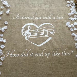 May include: A burlap aisle runner with white flower petals on the sides. The runner has a white heart with musical notes and the text "It started out with a kiss. How did it end up like this?"