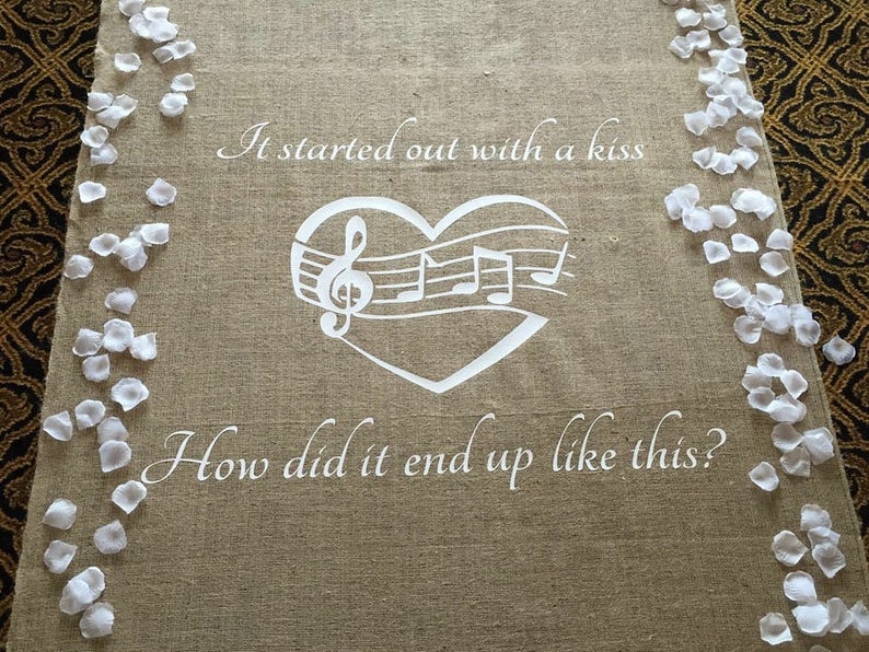 May include: A burlap aisle runner with white flower petals on the sides. The runner has a white heart with musical notes and the text "It started out with a kiss. How did it end up like this?"