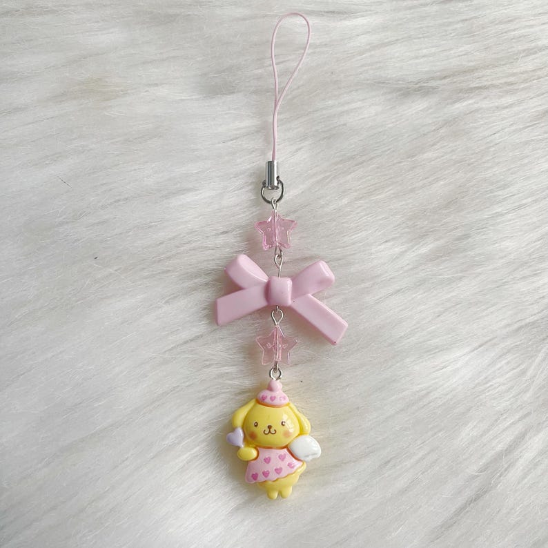 May include: A pink and yellow charm with a cartoon dog wearing a pink and white dress and holding a pink heart. The charm is attached to a pink ribbon and a string of pink stars.