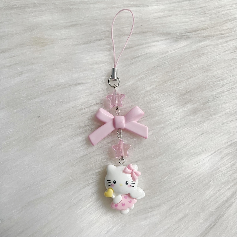 May include: Pink and white Hello Kitty phone charm with a pink bow and a yellow heart. The charm is attached to a pink string with pink star beads.