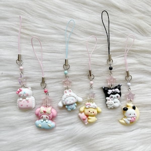 May include: Six cartoon character phone charms in various colours. Each charm features a different design, including a cat, bunny, and dog, with star and bead accents. The charms are attached to pink, blue, and black lanyards.