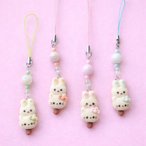 May include: Four pastel bunny-shaped charms on pink cords. Each charm has a different coloured cord and bead accents. The bunny charms have a popsicle stick base and a small flower or bow detail. The background is a solid pink.
