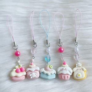Könnte beinhalten: Fünf bunte Handy-Anhänger in Cupcake-Form mit rosa, blauen und gelben Akzenten. Jeder Anhänger hat ein anderes Design, darunter Kirschen, ein Bär und eine Blume. Die Anhänger sind an rosa und blauen Lanyards mit silberfarbener Hardware befestigt.