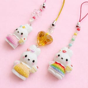 May include: Three white bunny charms with colorful outfits and accessories. One bunny wears a pink dress, another a yellow top, and the third a rainbow-striped outfit. Each charm has a different colored cord and bead accents, with a heart-shaped charm on one.