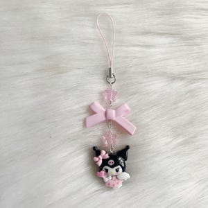May include: A pink and black character charm with a pink bow and pink stars hanging from a pink string. The character is wearing a pink dress and has black hair and a black face.