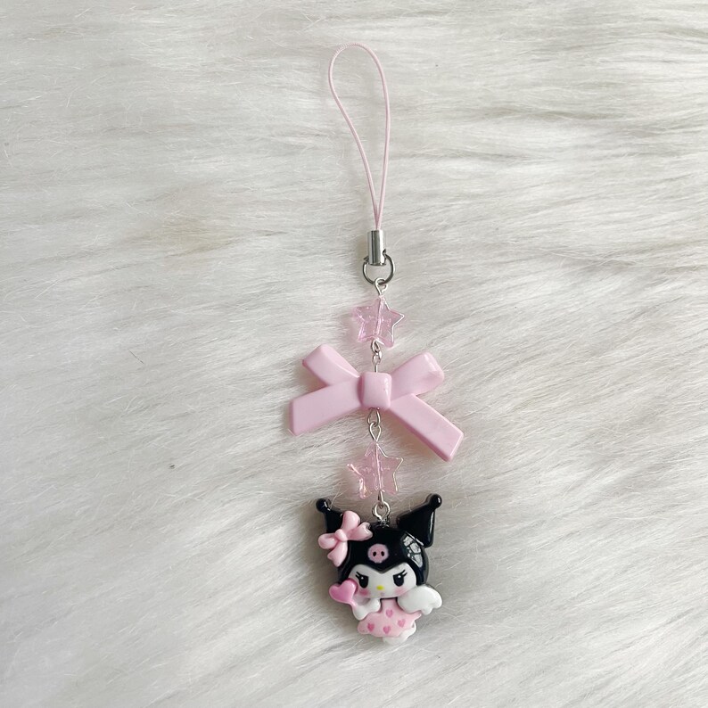 May include: A pink and black character charm with a pink bow and pink stars hanging from a pink string. The character is wearing a pink dress and has black hair and a black face.