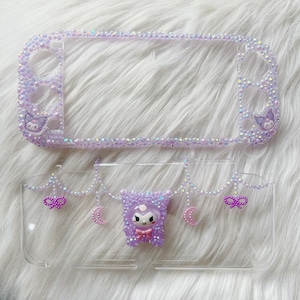 May include: A clear plastic case with a purple and white design. The case is decorated with rhinestones and features a small purple and white character with a bow. The case is designed to fit a Nintendo Switch.