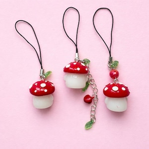 May include: Three mushroom-shaped charms with red caps and white spots, white stems, and black cords. One charm has a chain with red beads and green leaves. The charms are on a pink background.