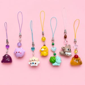 Puede incluir: Siete colgantes coloridos para teléfono con varias figuras pequeñas de plástico de animales y criaturas. Cada colgante incluye un lazo de cordón, herrajes de metal y cuentas decorativas con formas de corazones, estrellas y esferas.