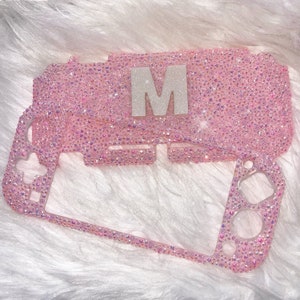 May include: Pink and iridescent glitter case for a Nintendo Switch console with the letter "M" in white glitter on the top.