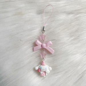 May include: A pink and white phone charm featuring a cartoon dog with a pink bow and a pink star charm.