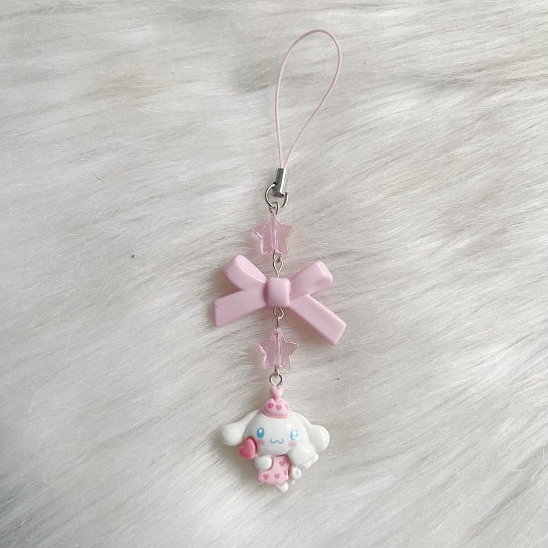 May include: A pink and white phone charm featuring a cartoon dog with a pink bow and a pink star charm.