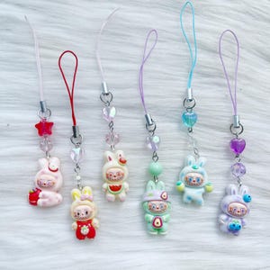 May include: A selection of colourful bunny-shaped phone charms. Each charm has a unique design, including strawberries, watermelon, and blueberries. The charms are attached to a string with beads and a metal clasp.