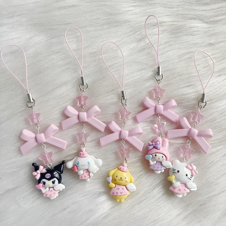 May include: Six pink and white phone charms with different cartoon characters. Each charm features a pink bow and a star-shaped charm. The characters include a black and white cat, a yellow dog, a pink and white bunny, a pink and white bear, a pink and white cat, and a pink and white unicorn.
