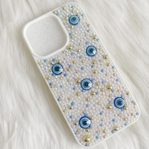 Op de afbeelding: Witte iPhone-hoes met een glinsterende, regenboogkleurige achtergrond en blauwe boze oog-charms.