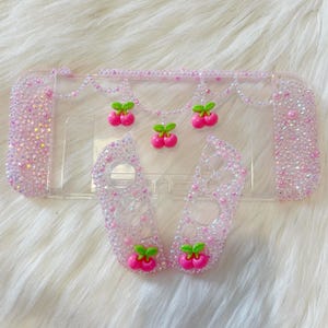 May include: Clear plastic Nintendo Switch case with a pink and white cherry design. The case is covered in rhinestones and has a dangling cherry charm.