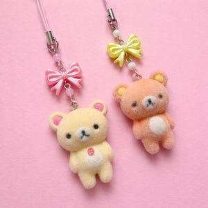 May include: Two plush bear charms in yellow and peach, each with a bow and a pink lanyard. The yellow bear has a red button on its chest. The charms are on a pink background.