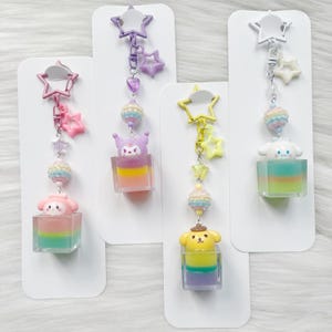 Op de afbeelding: Vier kleurrijke bedel sleutelhangers met stripfiguren. Elke sleutelhanger heeft een stervormige bovenkant, een kralen gedeelte en een karakter in een doorzichtige kubus. De karakters zijn onder andere My Melody, Kuromi, Cinnamoroll en Pompompurin.
