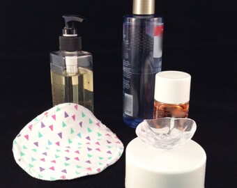 May include: A set of three clear bottles with pumps and lids, a white cotton round with a pink, purple, and green triangle pattern, and a white plastic container with a clear plastic lid.