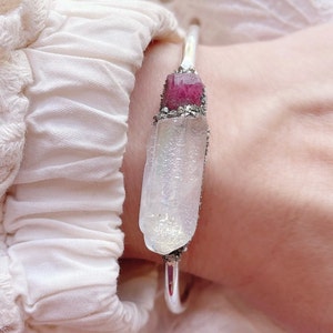May include: A silver-toned bracelet featuring a large, clear crystal with a pink crystal accent. The crystals are set with a textured, silver-colored metal. The bracelet is shown on a wrist, with a white fabric background.