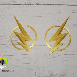 May include: Two gold-colored The Flash logos, each with a lightning bolt inside a circle, are displayed on a white wooden surface. The logos are 3D printed and have a textured finish.