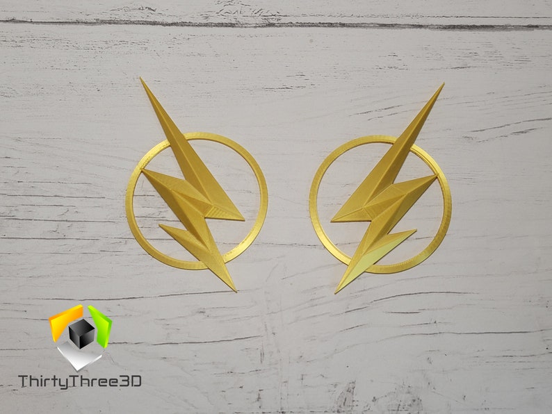 May include: Two gold-colored The Flash logos, each with a lightning bolt inside a circle, are displayed on a white wooden surface. The logos are 3D printed and have a textured finish.