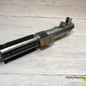 May include: A silver lightsaber prop with a black grip and a red button. The lightsaber has a gold detail on the side with a series of numbers.