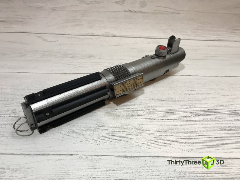May include: A silver lightsaber prop with a black grip and a red button. The lightsaber has a gold detail on the side with a series of numbers.