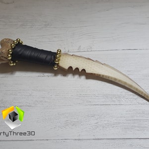 May include: A white bone-like dagger with a black leather handle and gold beads. The dagger has a serrated edge and a curved blade.