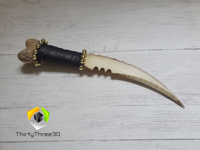 May include: A white bone-like dagger with a black leather handle and gold beads. The dagger has a serrated edge and a curved blade.