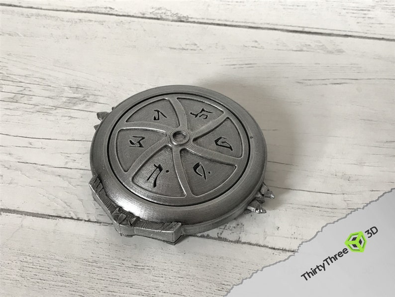 May include: A silver-colored metal disc with a circular design and six sections. Each section has a different symbol or design. The disc has a raised edge and a small hole in the center.