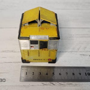 May include: A yellow, weathered, miniature model of a building with a black roof and windows. The model is sitting on a wooden surface with a ruler for scale.