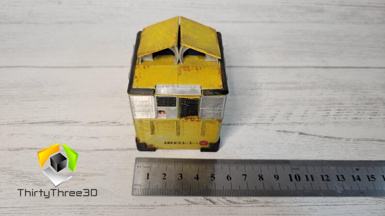 May include: A yellow, weathered, miniature model of a building with a black roof and windows. The model is sitting on a wooden surface with a ruler for scale.