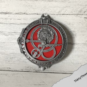 May include: A silver-coloured metal pendant with a red circular design in the centre. The pendant features intricate details and a circular inscription around the edge.