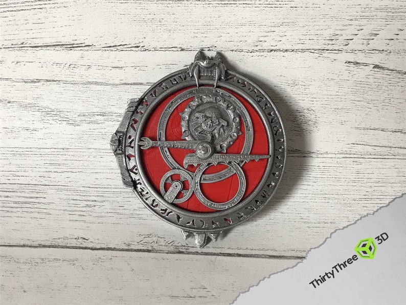 May include: A silver-coloured metal pendant with a red circular design in the centre. The pendant features intricate details and a circular inscription around the edge.