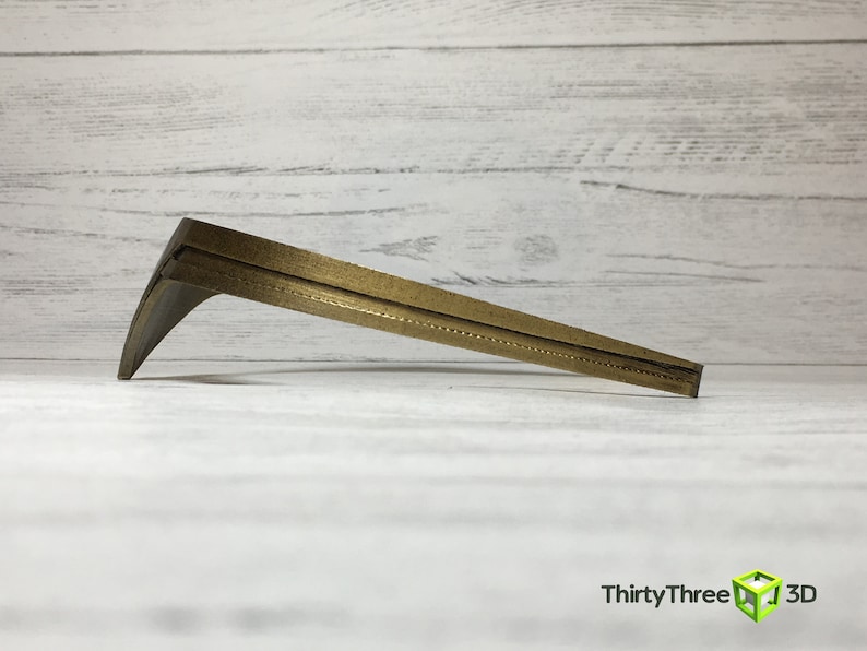 May include: A gold-colored metal bracket with a curved shape and a textured surface. The bracket is designed to support a shelf or other object.