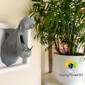 May include: A gray, 3D printed, geometric hippopotamus head sculpture in a white frame. The sculpture is mounted on a wall with a plant in a white pot in the background.