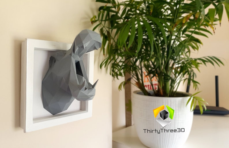 May include: A gray, 3D printed, geometric hippopotamus head sculpture in a white frame. The sculpture is mounted on a wall with a plant in a white pot in the background.
