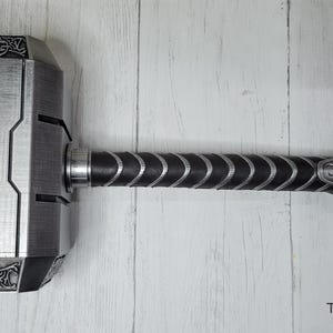 May include: A silver-colored Mjolnir hammer replica with a black leather handle and a strap. The hammer head has intricate details and a textured surface.