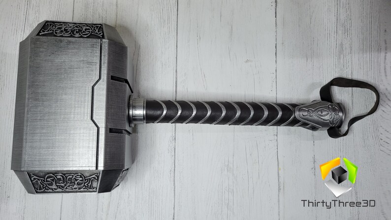 May include: A silver-colored Mjolnir hammer replica with a black leather handle and a strap. The hammer head has intricate details and a textured surface.