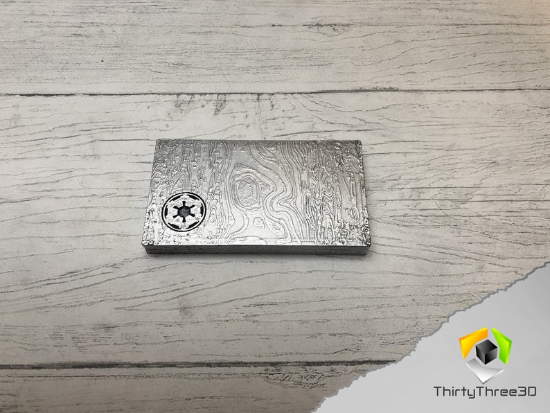 May include: A silver metal plate with a raised, textured design. The plate features a circular cutout in the center with a star-shaped symbol inside.