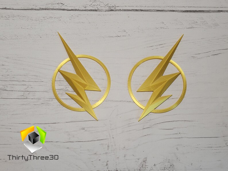 Op de afbeelding: Twee goudkleurige The Flash-logo's, elk met een bliksem in een cirkel, worden weergegeven op een wit houten oppervlak. De logo's zijn 3D-geprint en hebben een textuur.