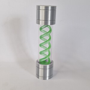 May include: A cylindrical glass vial with a green, spiraling design inside. The vial is capped with brushed metal on both ends. The design resembles a DNA strand. The vial is standing upright.
