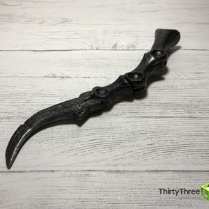 May include: A black, curved, claw-like object with a textured surface. The handle has a series of circular indentations. The object is set against a white wooden background.