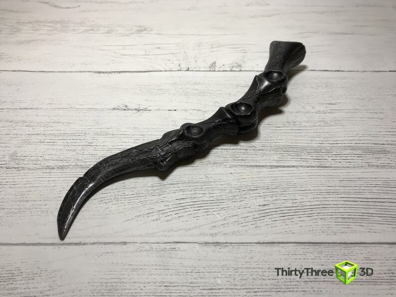 May include: A black, curved, claw-like object with a textured surface. The handle has a series of circular indentations. The object is set against a white wooden background.