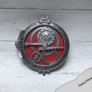 May include: A silver metal circular pendant with a red center and intricate gears. The pendant has the text "DOOM OF GUNMAR ECLIPSE IS MINE TO COMMAND" around the edge.