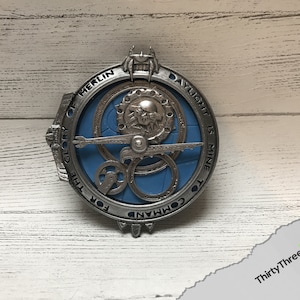 May include: A silver-colored metal pendant with a blue center and intricate gears. The text "FOR THE GLORY OF MERLIN DAYLIGHT IS MINE TO COMMAND" is inscribed around the edge.