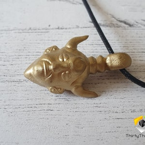 May include: Gold-colored pendant necklace featuring a detailed, stylized face with horns. The pendant is attached to a black cord.